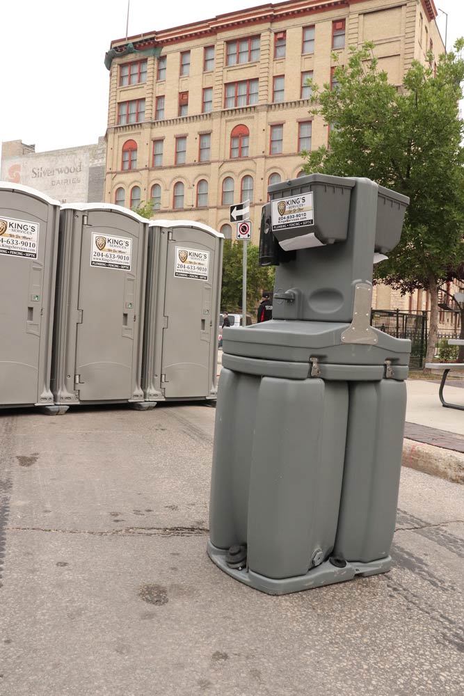 Row of gray portable toilets and a handwashing station on a city street. Brick building in background.