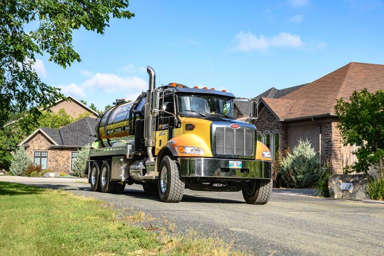 Yellow and black septic tank truck driving on a paved driveway in front of a house.