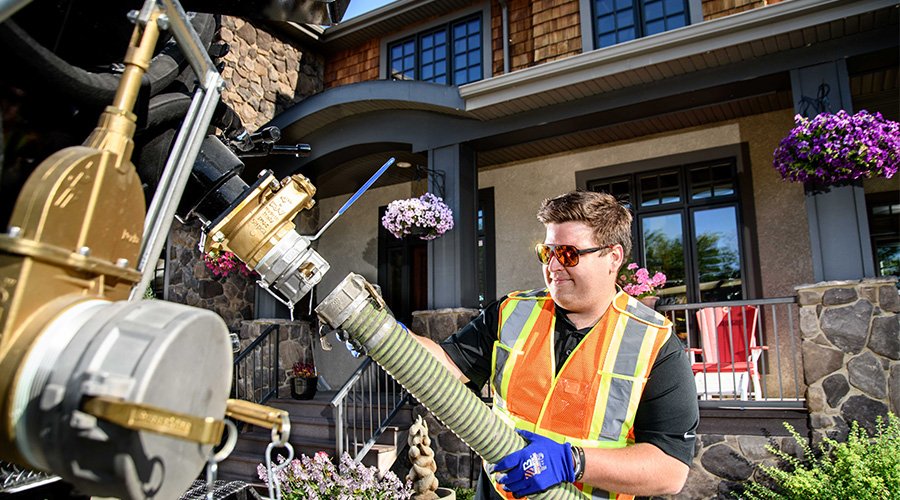 Fuel truck worker connecting hose to a house tank; orange vest, protective eyewear, sunny day.