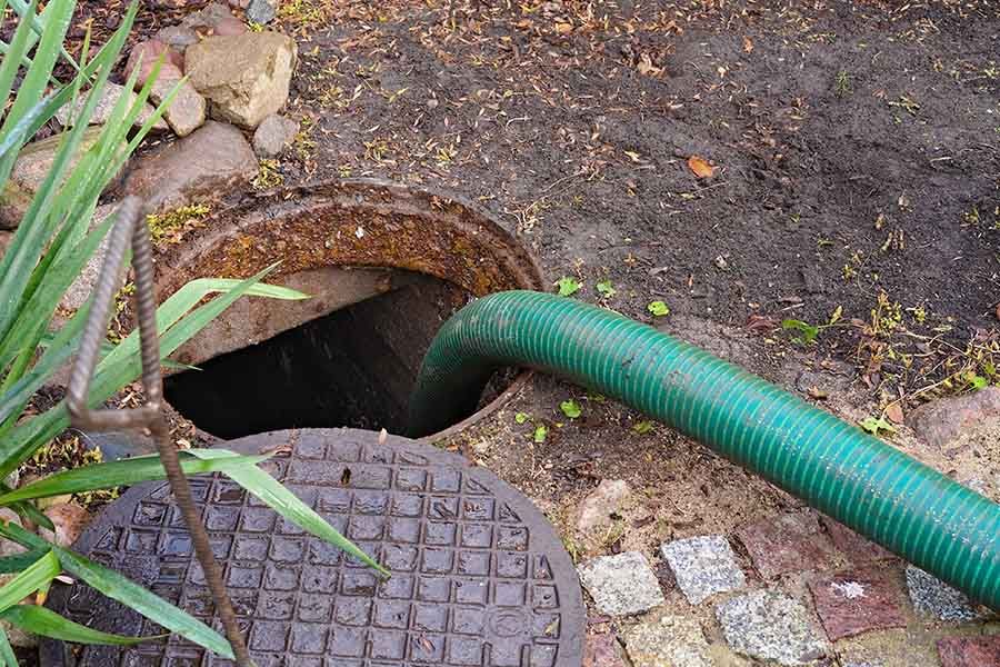 Green hose feeding into a round underground drain opening beside a manhole cover on dirt and paving stones